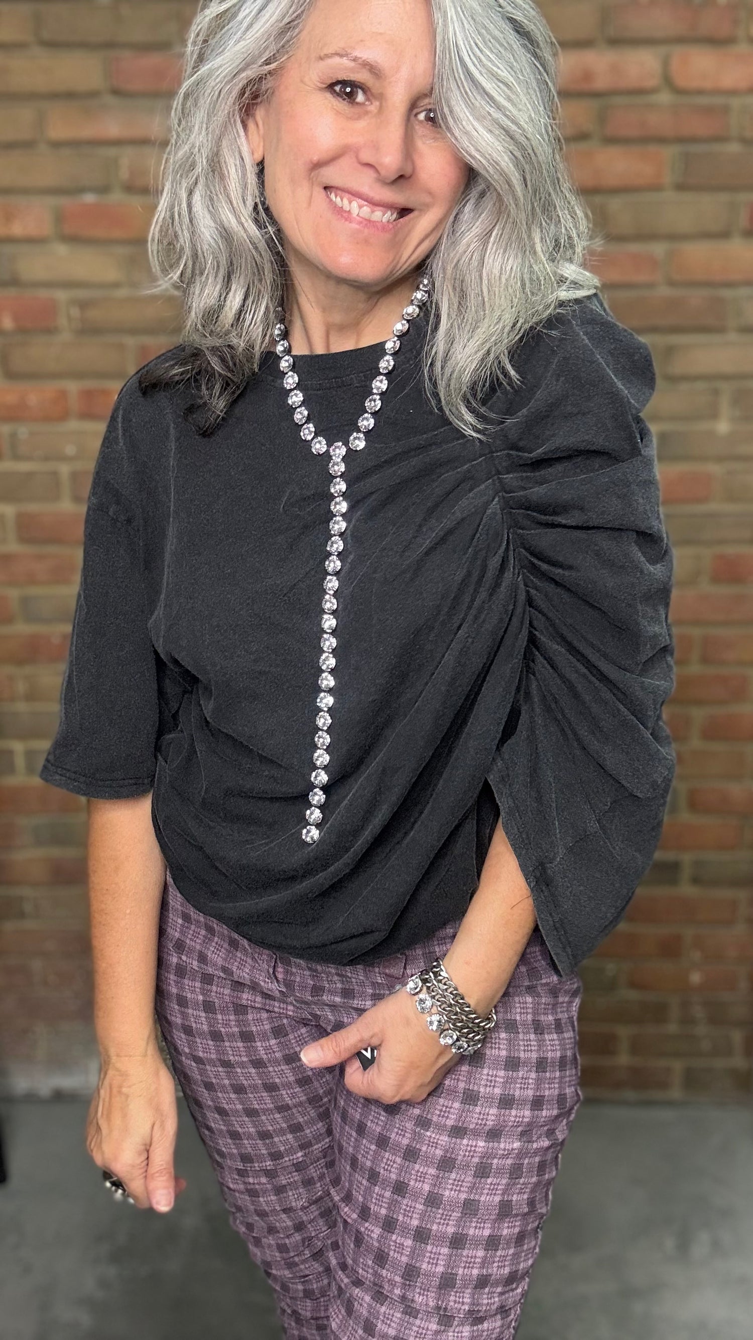 Woman wearing a black top with pearl details and plaid pants against a brick wall.