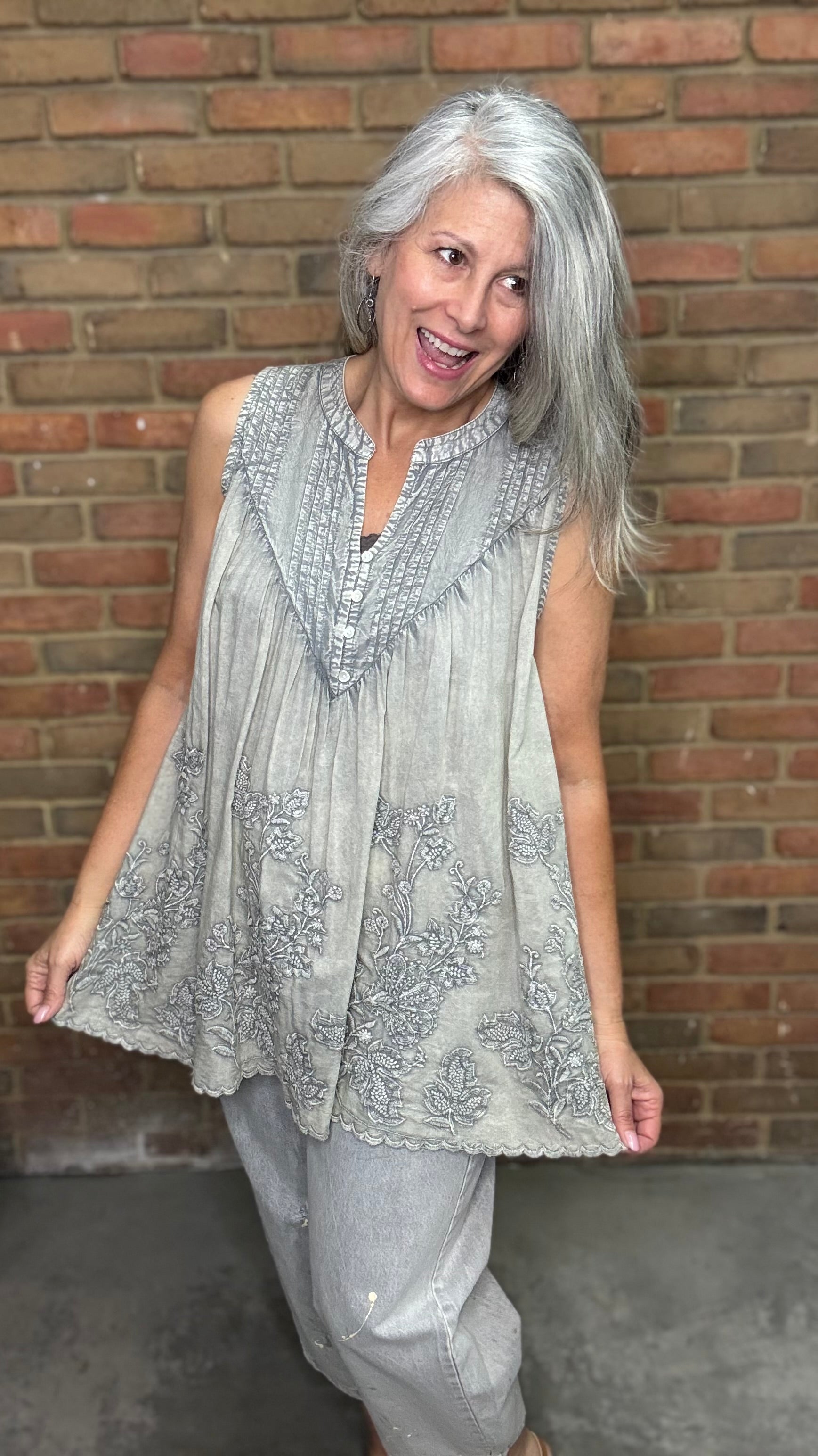 Woman wearing the Sage Floral Embroidery Tank Top, a light-colored sleeveless top with a pattern against a brick wall.