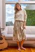 Woman in a cozy room wearing a cable knit sweater and floral dress, with a guitar and couch in the background.